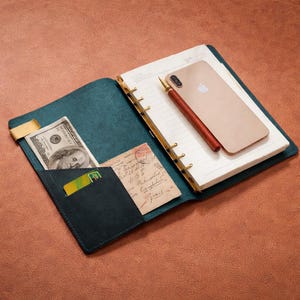 May include: Open teal leather journal with a gold-colored ring binder. Inside, there's a notepad, a gold-colored pen, and a smartphone. A pocket holds a $100 bill, a credit card, and a vintage postcard. The journal is open on a brown surface.