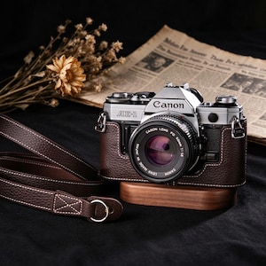 Pode incluir: Uma câmara de película Canon AE-1 prateada de 35 mm com lente preta, inserida numa meia capa de couro castanho texturado. A câmara inclui uma correia de pescoço em couro castanho a condizer com costuras brancas e repousa sobre uma base de madeira.
