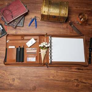 May include: A tan leather zippered portfolio organizer featuring a metal ring binder with white paper, multiple card slots, a pen holder, and a zippered pocket. A small paper cone filled with white artificial flowers is tucked inside.