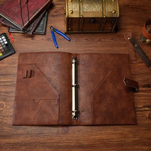 May include: A brown leather binder featuring a central three-ring metal mechanism, interior pockets for documents, and two pen loops. The binder includes a side strap closure with a snap button, providing a secure way to organize papers.