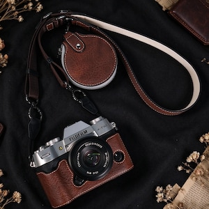 May include: A silver Fujifilm X-T30 camera featuring a brown leather-textured protective case and a matching brown leather camera strap with an attached circular brown leather pouch. The lens is black with white text detailing the optical specifications.