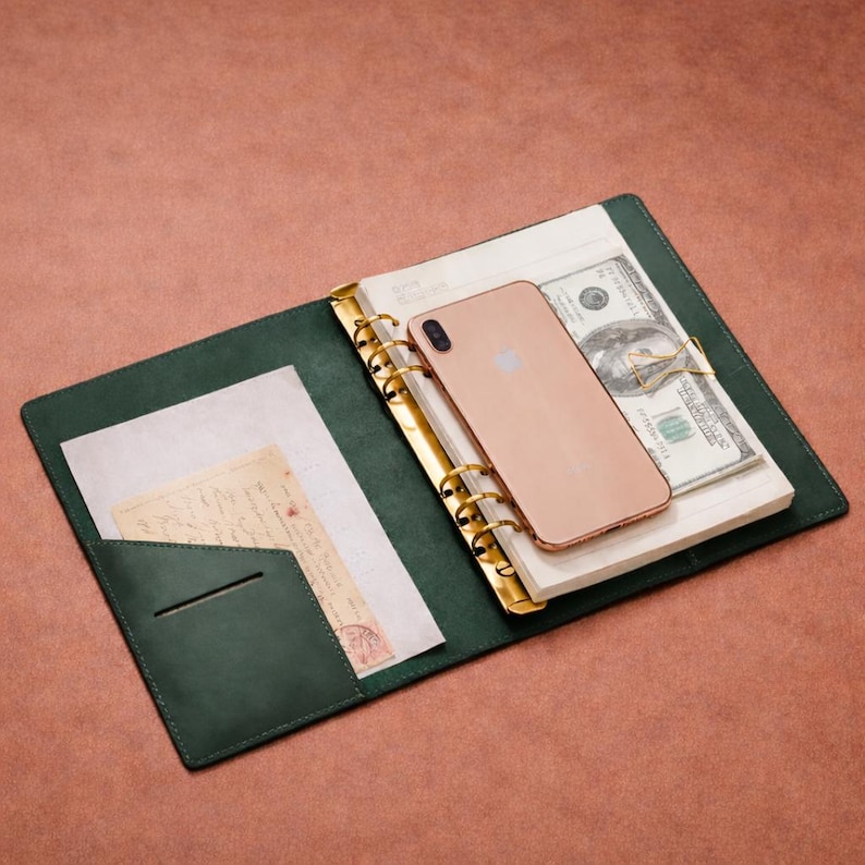 May include: Open green leather binder with gold rings, holding a stack of paper, a smartphone, and a wad of US hundred-dollar bills. A pocket holds a piece of paper and a vintage postcard. The binder is open on a brown surface.