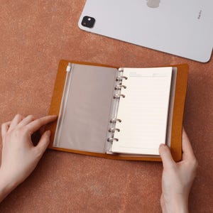 Könnte beinhalten: Offenes, hellbraunes Leder-Notizbuch mit linierten Seiten und transparenten Kunststofftaschen. Das Notizbuch wird von zwei Händen offen gehalten. Ein silbernes Tablet befindet sich im Hintergrund.