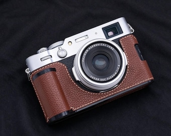 May include: A silver digital camera with a black textured grip and a brown leather case. The lens is labeled Fujinon Aspherical Lens Super EBC f=23mm 1:2 II. The camera features a silver top plate with a shutter button and a viewfinder window.