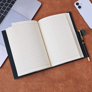 Könnte beinhalten: Ein offenes Notizbuch mit linierten Seiten und einem dunkelgrünen Einband, zusammen mit einem schwarzen Stift mit goldenen Akzenten. Ein Laptop und ein Smartphone befinden sich im Hintergrund auf einer braunen Oberfläche. Das Notizbuch ist schreibbereit.
