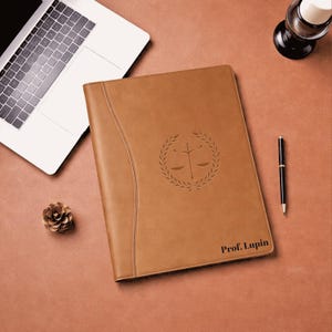 May include: A tan leather portfolio with a debossed scales of justice emblem and the name "Prof. Lupin." The portfolio is on a brown surface with a laptop, pen, pinecone, and desk lamp.