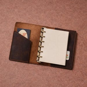 May include: Open leather journal with a brown cover and a spiral binding. The journal has a small pocket on the left side and a card in the pocket. The journal is open to a blank page. A small card is visible on the right side.