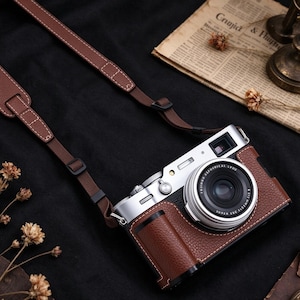 May include: A silver digital camera with a brown textured leather case and a matching brown adjustable camera strap. The lens is labeled FUJINON ASPHERICAL LENS SUPER EBC XF 23mm 1:2 R. The camera features a silver top plate and a black grip area.