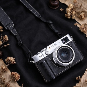 May include: A silver and black digital camera featuring a textured black leather-like protective case and a matching black camera strap with white stitching. The lens is labeled Fujifilm Super EBC 1:2 23mm, and the camera body has a silver top plate.