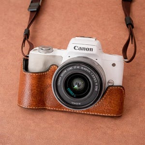 May include: A white Canon camera with a black lens is in a brown leather case. The camera has a brown strap and is on a brown background. The Canon logo is visible on the camera body.