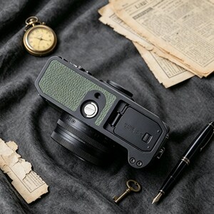 May include: A digital camera featuring a textured olive green leatherette grip on its black body, positioned alongside a gold pocket watch, aged paper documents, a small brass key, and a black fountain pen on a dark gray fabric surface.