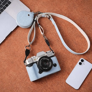 Könnte beinhalten: Hellblaue Fujifilm X-Too Kamera mit passendem Gehäuse und Riemen. Die Kamera hat ein schwarzes Objektiv und silberne Akzente. Ein kleines, rundes, hellblaues Etui und ein weißes Smartphone sind ebenfalls sichtbar.