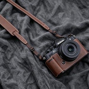 May include: A black Nikon Zf camera featuring a brown textured leather-like half case and a matching brown leather camera strap with white stitching. The camera body includes a lens, a top-mounted dial, and the Nikon Zf logo clearly visible on the front.