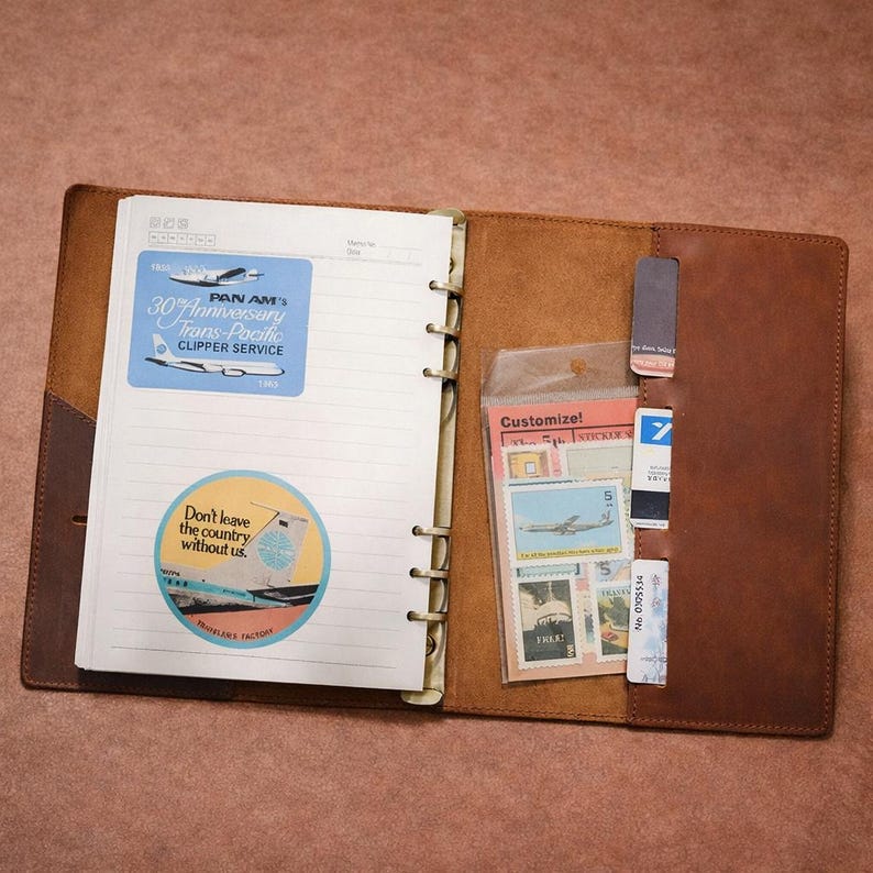 May include: Open leather journal with lined pages, featuring Pan Am stickers and a clear plastic sleeve with stamps. The journal includes a sticker that says "Don't leave the country without us."