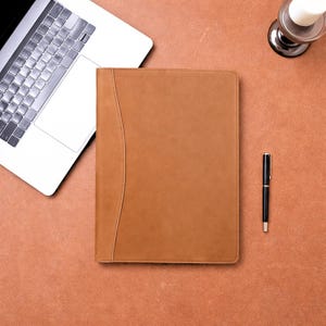 May include: A tan leather portfolio with a curved design detail, a black pen, and a laptop are arranged on a brown surface. A candle holder is also visible. The items are arranged for a workspace or office setting.