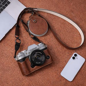 Könnte beinhalten: Eine braune Leder-Kameratasche mit passendem Objektivdeckel und Riemen. Die Kamera ist silber und schwarz, mit dem Markennamen "FUJIFILM" sichtbar. Ein weißes Smartphone befindet sich ebenfalls im Bild.