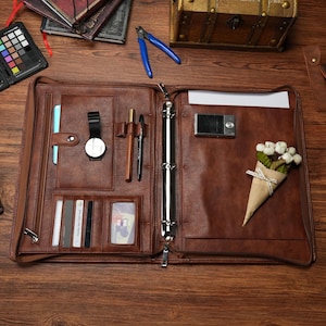 May include: A brown leather portfolio organizer featuring a central three-ring binder mechanism, multiple card slots, a pen holder, a zippered pocket, and a dedicated sleeve for a tablet. The interior includes a small bouquet of white flowers.