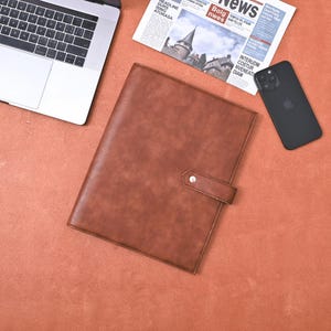 May include: A brown leather portfolio with a strap closure and silver button. A laptop, newspaper, and black smartphone are on a reddish-brown surface. The newspaper has the headline "News".