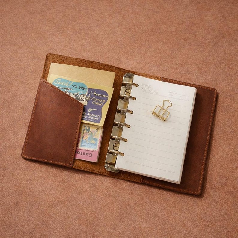 May include: Open brown leather journal with a ring binder, a small pocket, and a gold binder clip. The pocket contains vintage-style cards and stamps. The journal is open to a blank page with lines.