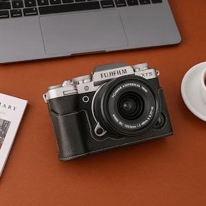 May include: A Fujifilm XTS camera with a black leather case sits on a brown surface, next to a laptop and a cup of coffee. The camera lens is prominent, with the brand name visible. The case has white stitching.