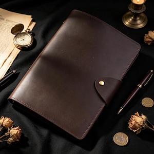 May include: A dark brown leather notebook cover featuring a smooth finish, rounded corners, and a single brass snap button closure. The rectangular cover is constructed with visible stitching along the edges, providing a structured and durable appearance.