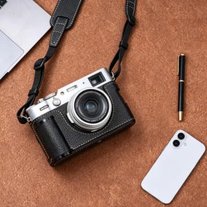 Pode incluir: Câmera de couro preta com lente prateada e alça preta. A câmera está em uma superfície marrom, ao lado de um smartphone branco, uma caneta preta e um laptop.