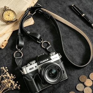 May include: A black leather camera strap with silver-toned metal rings and stitching, attached to a silver and black Fujifilm X-T30 camera. The strap features a smooth black exterior and a light-colored interior lining, designed for carrying the camera.
