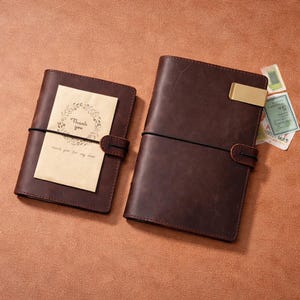 May include: Two brown leather journals with elastic closures. One journal has a card that reads "Thank you" with a floral wreath design. The other journal has a gold-colored metal clip. Small decorative stamps are visible.