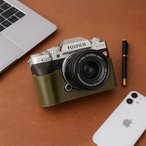 Könnte beinhalten: Eine silberne Fujifilm X-T5 Kamera mit schwarzem Objektiv und olivgrüner Lederhülle. Ein schwarzer Stift und ein weißes iPhone sind ebenfalls sichtbar. Die Kamera befindet sich auf einer braunen Oberfläche.