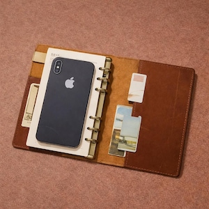 May include: Open brown leather binder with a black smartphone and several cards inside. The binder has a gold-colored ring mechanism and a cream-colored paper insert. The background is a solid, muted pink.