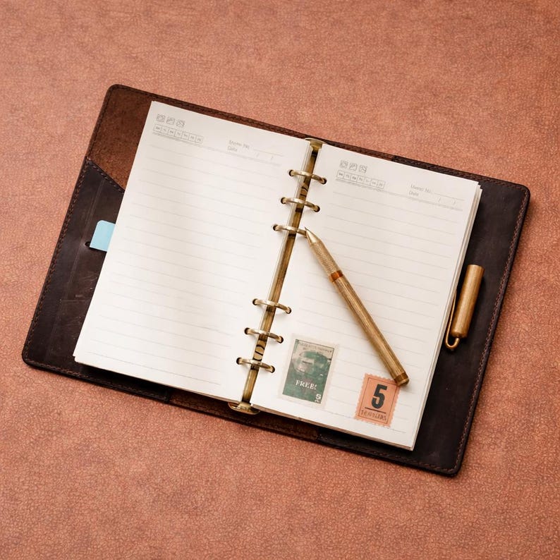 May include: Open leather-bound notebook with lined pages, a gold pen, and two postage stamps. The notebook is brown, with a dark brown cover and a light blue bookmark. The pen is gold with a brown accent. The stamps are green and orange.