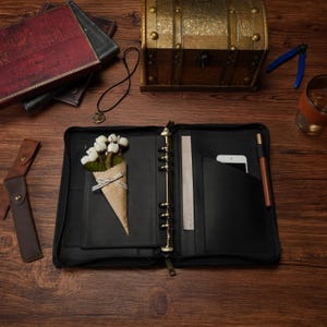 May include: A black leather zippered binder featuring a six-ring metal mechanism, a vertical pocket containing a white smartphone, a pen loop with a brown pen, and a decorative paper cone filled with white fabric flowers attached to the interior.