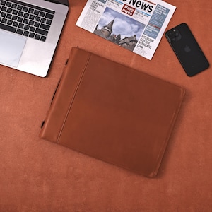 May include: A brown leather portfolio with a handle, a laptop, a newspaper, and a black smartphone are arranged on a reddish-brown surface. The newspaper has the headline "HEADLINE DURS ISING ATORASA".