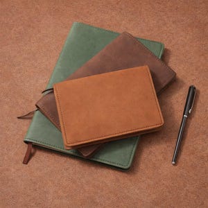 May include: A collection of leather journals and a pen. The journals are in shades of green, brown, and tan. The pen is black with silver accents. The journals are stacked on top of each other.