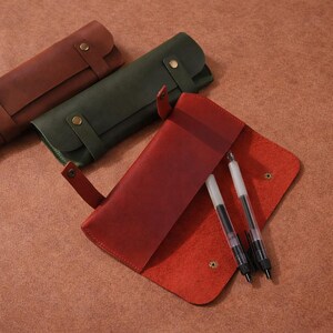 Op de afbeelding: Leren etuis in rood, groen en bruin. De rode etui is open en toont twee pennen. Elke etui heeft een klepsluiting met een knoop en een riem. De etuis liggen op een bruin oppervlak.