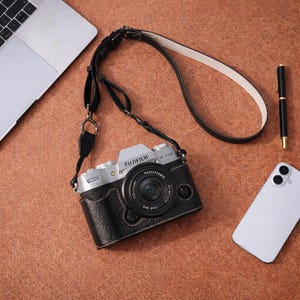 Könnte beinhalten: Eine silberne und schwarze Fujifilm X-T50 Kamera mit schwarzer Ledertasche und Riemen. Das Kameraobjektiv ist zentriert, und der Riemen ist über das Bild drapiert. Ein Laptop, ein Stift und ein Smartphone sind ebenfalls sichtbar.