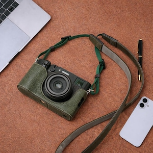 May include: A black digital camera with a green leather case and a green and gray strap. The camera is on a brown surface, with a laptop, pen, and smartphone nearby. The camera lens is centered.