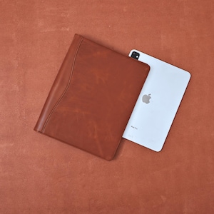 Könnte beinhalten: Ein braunes Leder-Portfolio mit geschwungenem Design-Detail, neben einem weißen iPad Pro. Das Portfolio hat eine glatte Textur und eine warme, rötlich-braune Farbe. Das iPad hat ein silbernes Apple-Logo.
