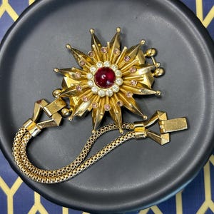 Pode incluir: Broche dourado com um design de estrela, apresentando uma pedra preciosa vermelha central rodeada por cristais transparentes e rosa. Uma delicada corrente e fecho dourados estão presos. O broche é exibido em um prato preto.