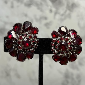 Op de afbeelding: Een paar vintage clip-on oorbellen. Elke oorbel heeft een bloemenontwerp met grote, facetgeslepen, dieprode edelstenen en kleinere rode stenen gezet in een zilverkleurig metaal. De oorbellen worden gepresenteerd op een zwarte standaard.
