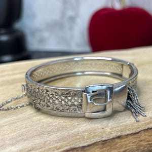 May include: Silver-toned hinged bracelet with intricate etched patterns and a buckle clasp. The bracelet features a decorative fringe of small chains. The jewelry is displayed on a wooden surface.