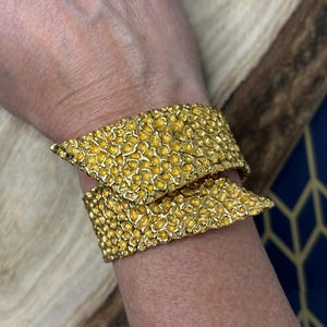 Könnte beinhalten: Ein goldfarbenes Armreif-Armband mit strukturierter Oberfläche und überlappendem Design. Das Armband hat ein unregelmäßiges, unebenes Muster. Der Schmuck ist ein Statement-Stück. Das Design des Armbands umschließt das Handgelenk.