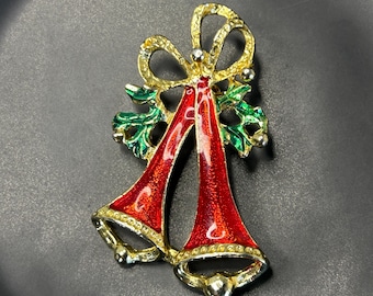 May include: A Christmas-themed brooch featuring two red bells with gold trim and a gold bow. Green holly leaves accent the bells. The brooch is set against a dark background.