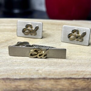 May include: A vintage set of accessories, including a tie clip and two cufflinks. The rectangular pieces are silver-toned with the gold-coloured word "Ed" in a cursive script. The set is displayed on a wooden surface.