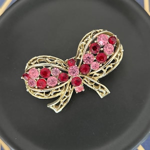 Könnte beinhalten: Goldfarbene Schleifenbrosche mit filigranem Design, verziert mit rosa und roten Strasssteinen. Die Brosche ist vor einem schwarzen Hintergrund platziert, der ihre komplizierten Details und lebendigen Farben hervorhebt. Ein Accessoire im Vintage-Stil.