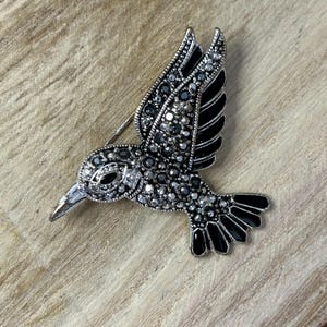 May include: A silver hummingbird brooch with black enamel accents and clear and black rhinestones.