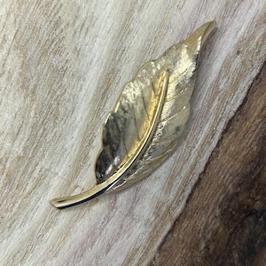 Op de afbeelding: Een gouden bladvormige broche. De broche is gemaakt van metaal en heeft een getextureerd oppervlak.