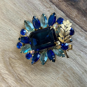 Może przedstawiać: Wintydżowa broszka z centralnym prostokątnym kamieniem w kolorze ciemnoniebieskim, otoczonym niebieskimi i turkusowymi kamieniami w kształcie łezki. Broszka ma złoty metalowy element i złoty liść.