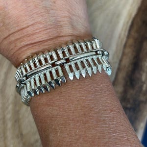 May include: A silver-toned metal bracelet featuring a repeating vertical spike or feather-like design with textured detailing. The piece has a central horizontal bar and a hinged construction, creating a structured, segmented appearance around the wrist.