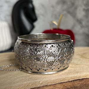 May include: Silver filigree bracelet with intricate floral and geometric cut-out designs. The bracelet is round with a small chain attached. The background includes a black chess piece and a red apple.